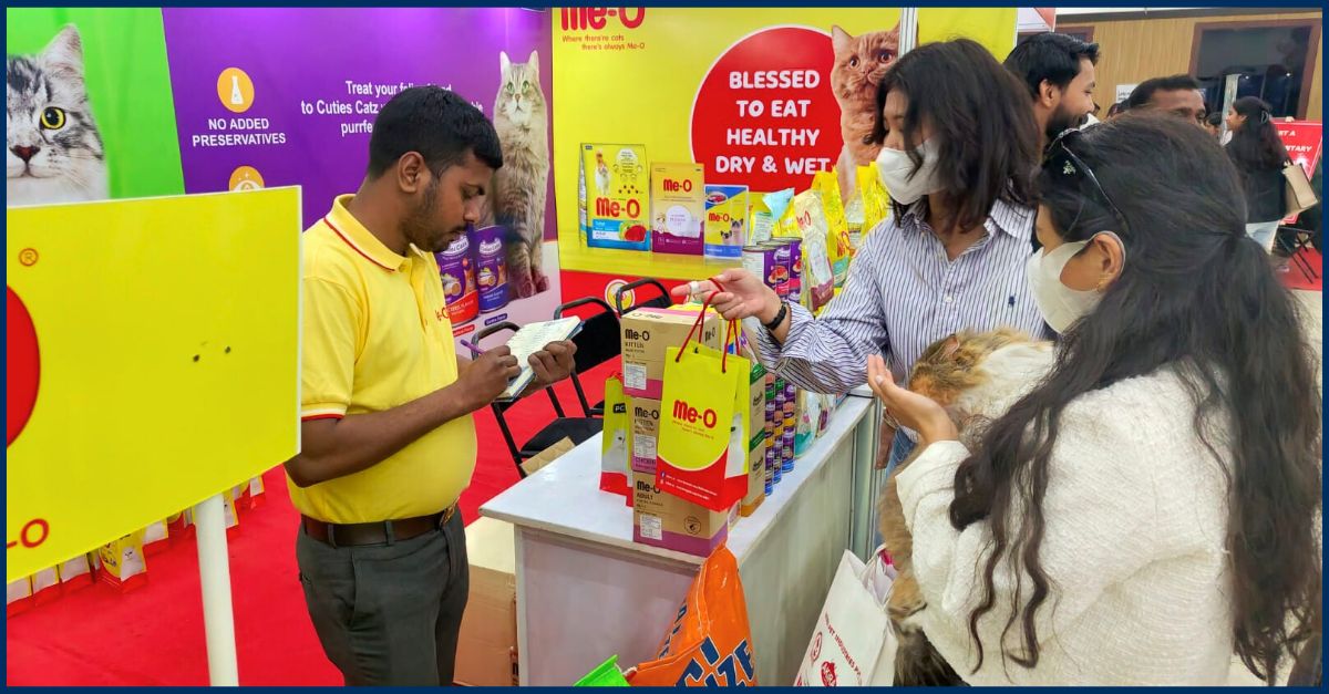 Me-O cat food brand representative in yellow polo demonstrating products at FCI Cat Show 2025 to visitors with live cat.